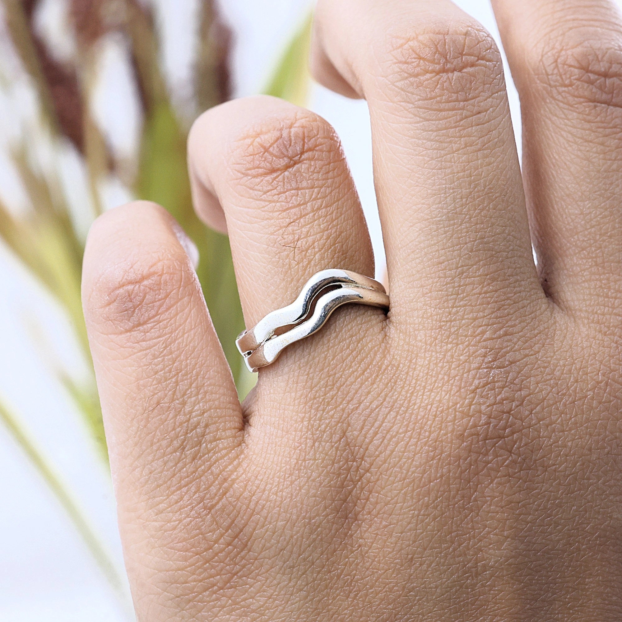 A close up side angle of a wave ring silver band ring with a unique double layered design worn on a finger in front of plants