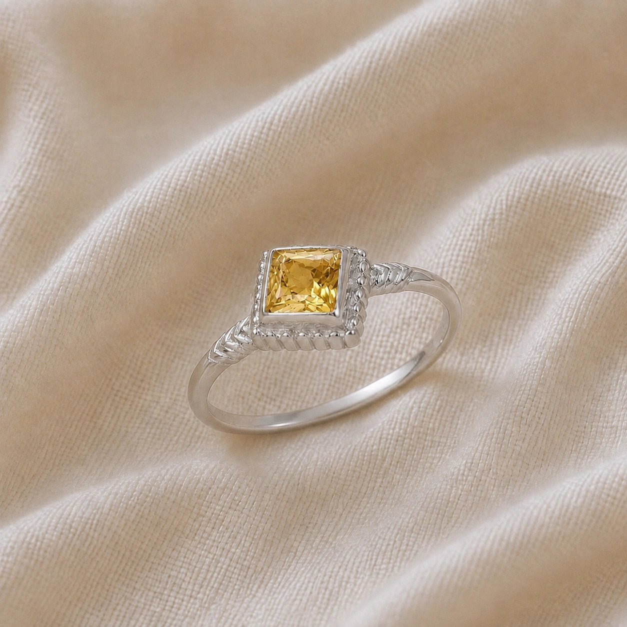 Princess_cut_ring_sterling_silver_with_square_yellow_sapphire_and_detailed_band_slightly_tilted_view_on_soft_fabric_background