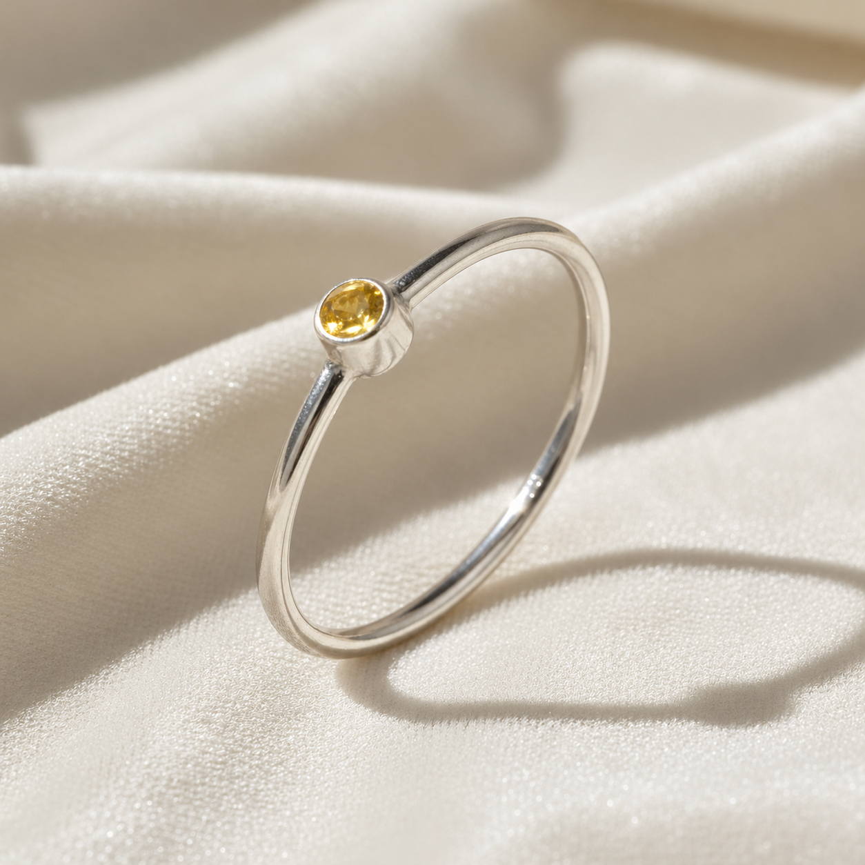 A sterling silver birthstone ring featuring a yellow sapphire gemstone, styled as a delicate stacking ring on a soft white fabric background