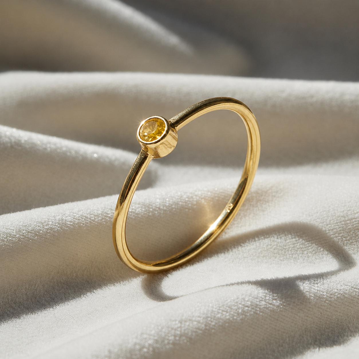 A gold plated birthstone ring featuring a yellow sapphire gemstone, styled as a delicate stacking ring on a soft white fabric background
