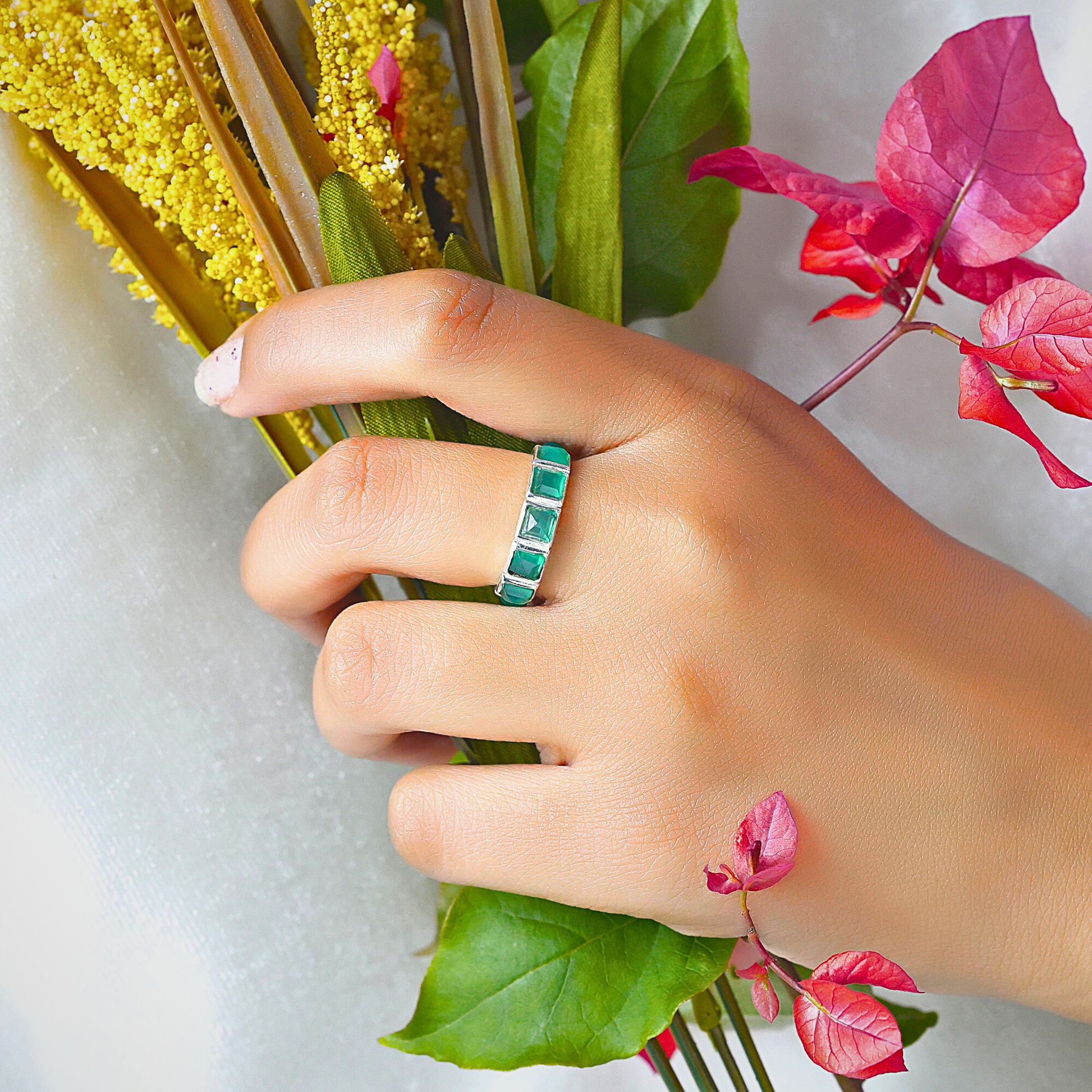 A hand worn a silver square-cut green onyx ring holding yellow and vibrant reddish-pink flowers.