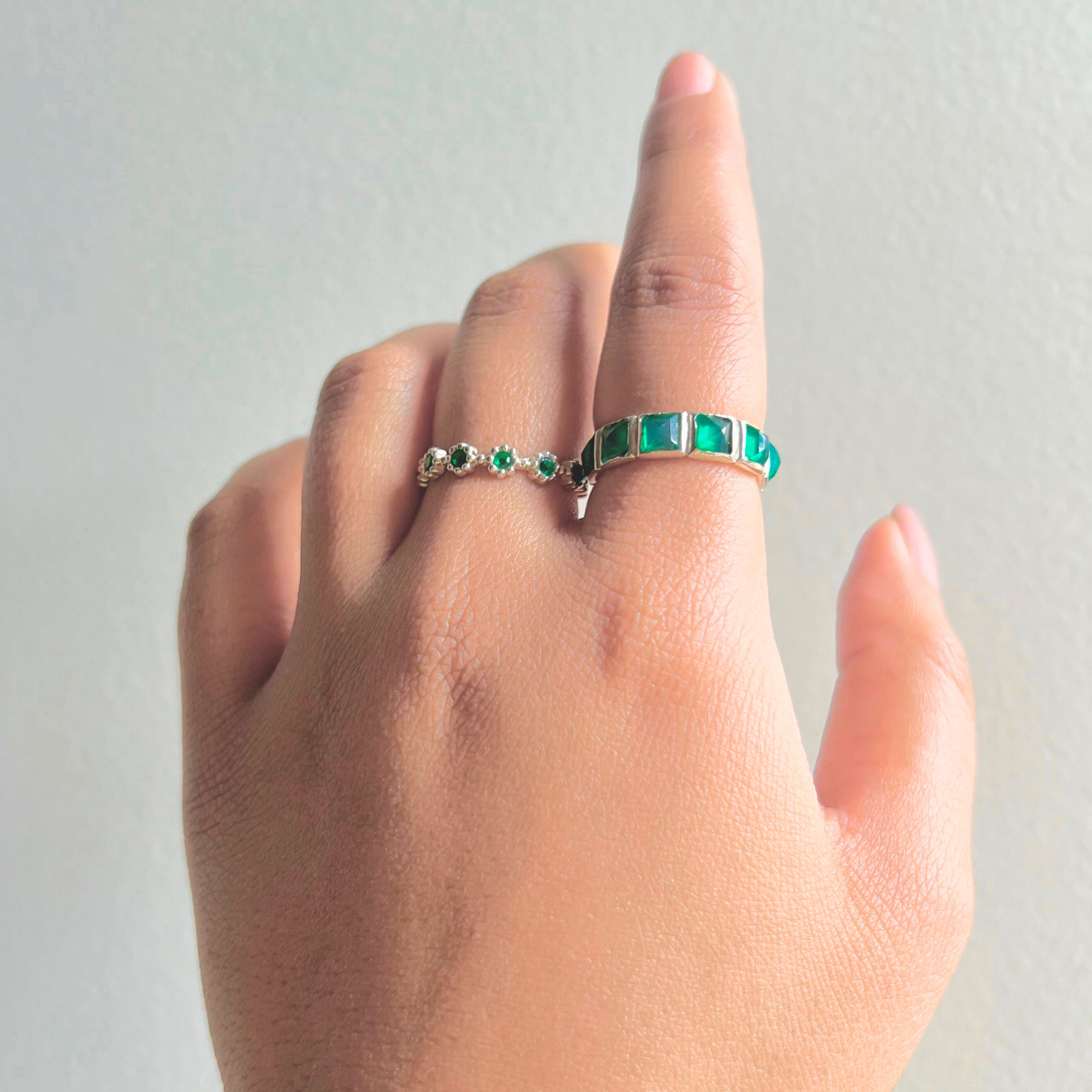 Green onyx ring worn on a model's finger alongside a small silver eternity band with tiny green stones against a plain gray wall background.