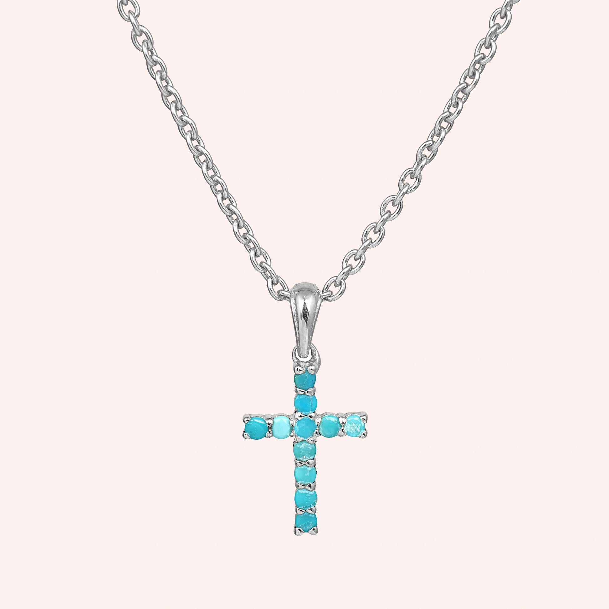 A sterling silver cross pendant necklace with turquoise stones shown in a front angle view over a light pink background