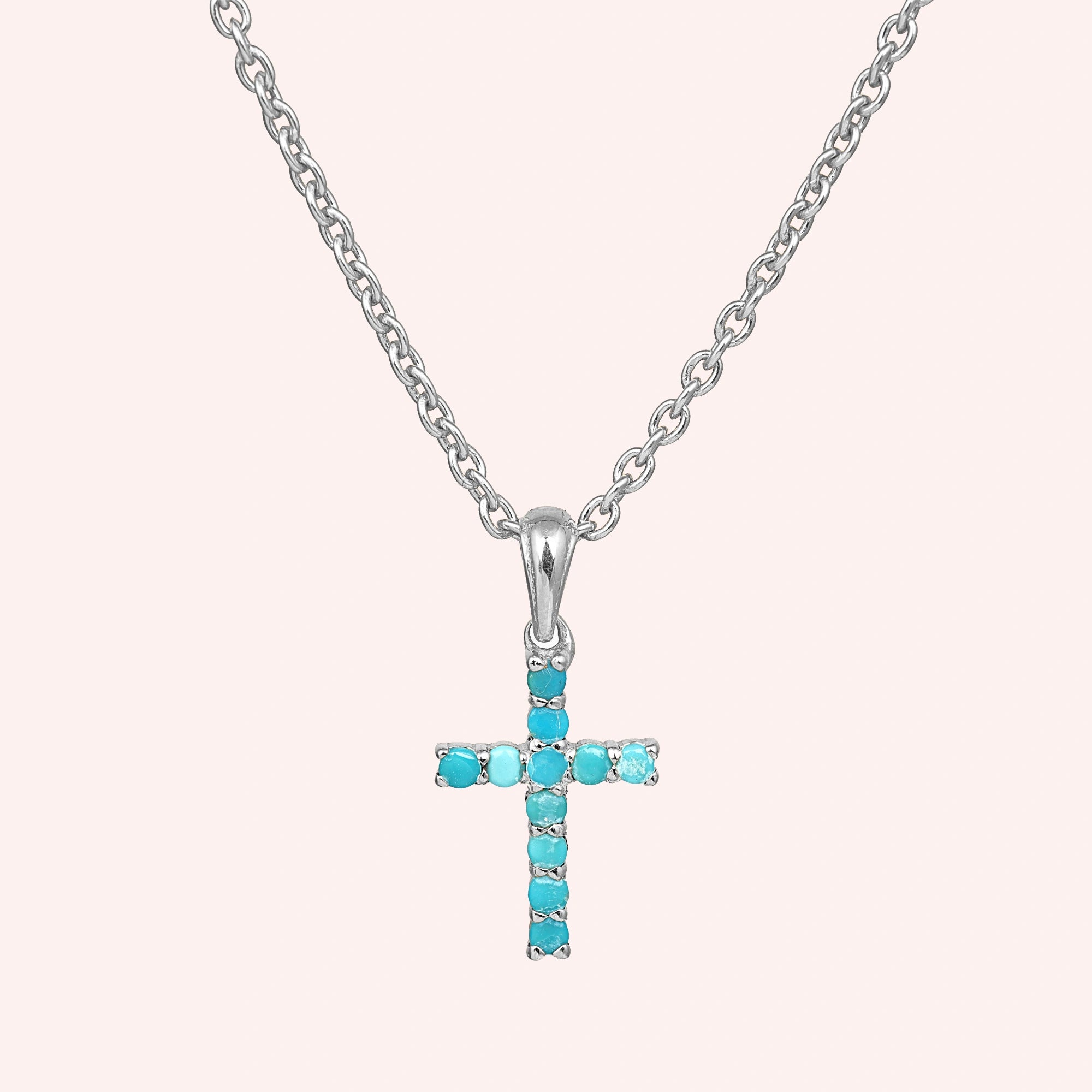 A sterling silver cross pendant necklace with turquoise stones shown in a front angle view over a light pink background